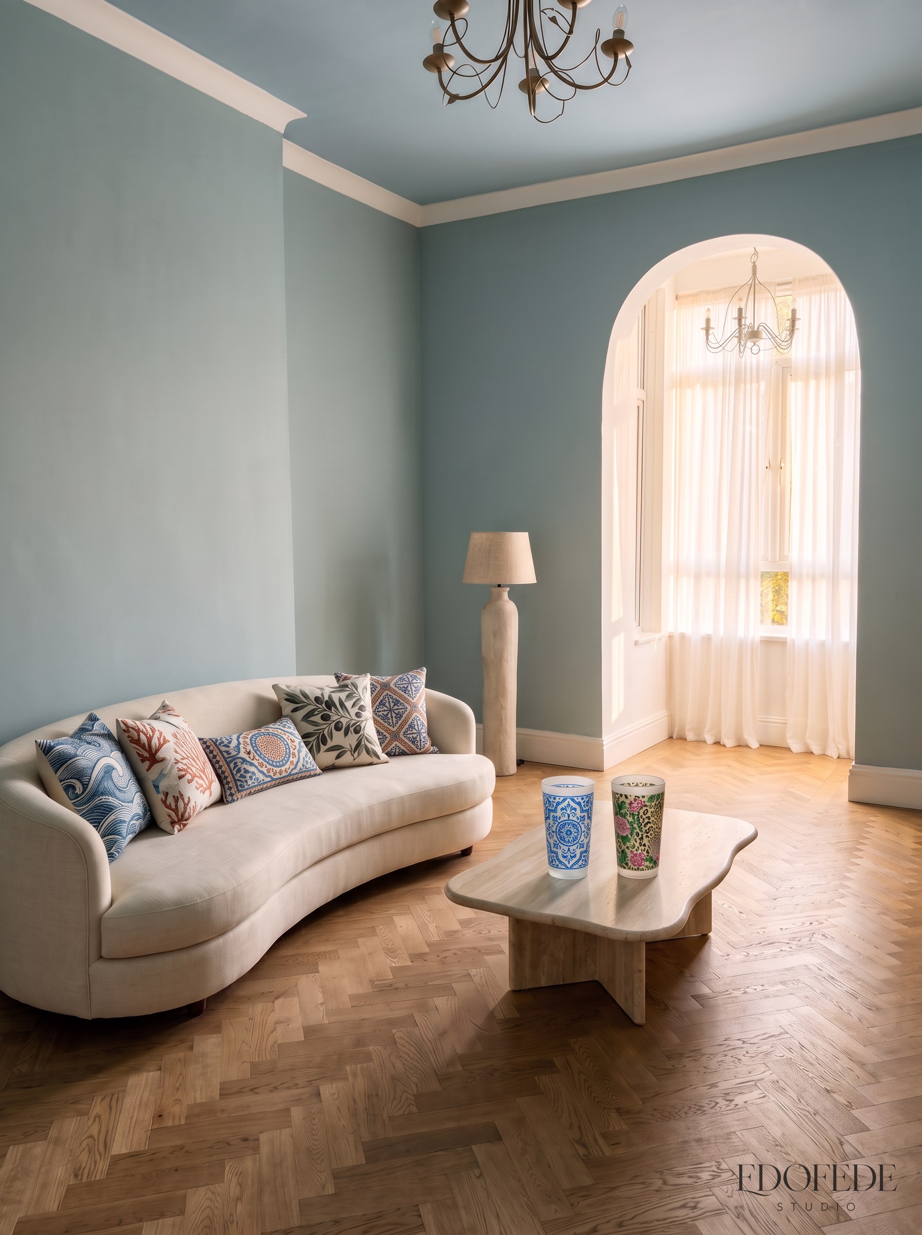 The Living Room concept: a powder blue ceiling above a cream curved sofa with Mediterranean cushions, two frosted patterned tumblers on a stone coffee table, and pale oak herringbone floor